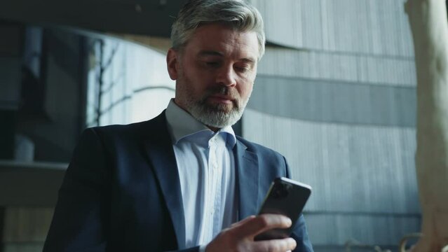 Close-up view of successful bearded man with gray hair using smartphone. Shot bearded handsome businessman smiling at camera on blurred background of business center. Indoor. Corporate, gadget. Busy