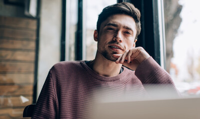 Portrait of handsome male blogger with cropped laptop computer looking at camera during time for doing distance job, skilled Caucasian hipster guy with blurred netbook posing in coworking space