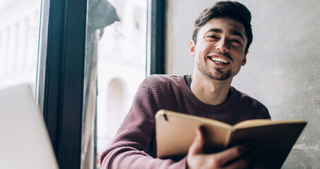 Portrait of happy Caucasian IT professional with modern laptop computer and education textbook smiling at camera, skilled male blogger with notepad and digital netbook posing in coworking space