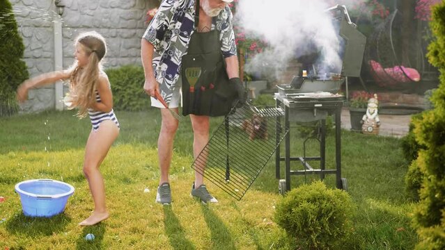 Old man is grilling in the backyard while children are splashing with water balloons on a hot summer day.