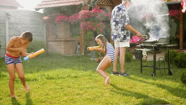 Two laughing kids splashing water with toy guns at backyard while while grandfather is grilling