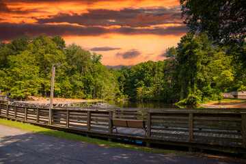 a gorgeous summer landscape in the park with a lake and a brown wooden bridge and a brown pergola surrounded by lush green trees, grass and plants with powerful clouds at sunset at Dupree Park - obrazy, fototapety, plakaty