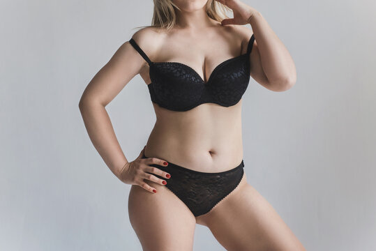 A young woman with curves and cellulite poses in black lingerie on a gray background without a face. Plus size model posing in lingerie. A plump blonde takes off her bra. The concept of body positivit