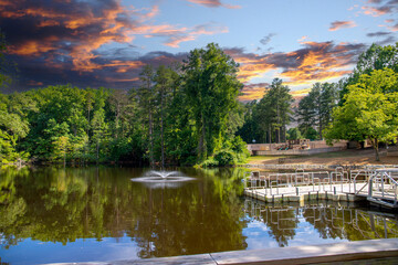 a long T shaped dock with a metal hand rail on the lake surrounded by lush green trees, grass and plants with powerful clouds at sunset at Dupree Park in Woodstock Georgia USA - obrazy, fototapety, plakaty