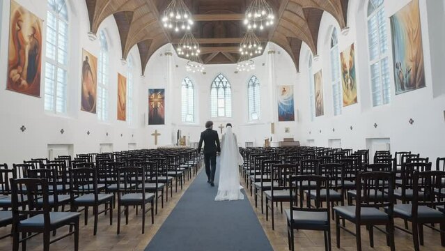Wedding ceremony just for bride and groom in chapel. Action. Rear view of a man in suit and a woman in white dress walking together along empty rows in church towards the altar.