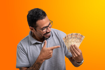man holds money, looks at the money in his hand smiles and points to the cedulas, Brazilian money, on orange background