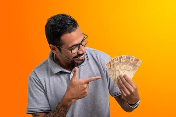 Fotobehang man holds money, looks at the money in his hand smiles and points to the cedulas, Brazilian money, on orange background © Edson Souza