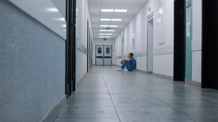 Stressed doctor sitting leaning on wall hallway. Tired medical worker resting.