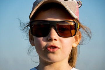 a little blonde girl in a pink baseball cap and big sunglasses makes a funny face and purses her lips in the bright sun