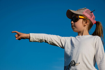 a little blonde girl in a pink baseball cap and large sunglasses stands in profile against a bright blue sky, purses her lips and points her finger into the distance