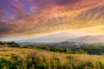 view from hill with golden grass and green bushes to a valley town with majectic mountains and scenic cloudy sunset on background