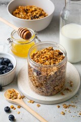 Healthy breakfast ingredients. Homemade granola with raisin, sunflower seeds, hazelnut and peanut in open glass jar, milk or yogurt bottle, honey and blueberries on grey background. Selective focus.