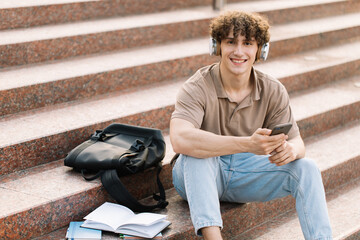 Attractive curly haired young man university or college student sits on the stairs and looking...