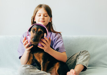 Happy teenage girl in lilac t-shirt sits on the sofa, listens to music has fun playing with the dog