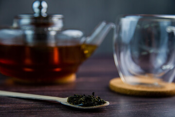 A wooden spoon with tea lies against a dark wood background, a mug and a teapot are visible from behind, a photo in dark colors. High quality photo