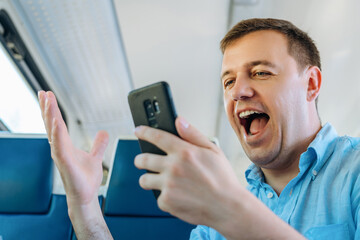 Overjoyed caucasian man looks at smartphone screen with mouth opened in train commuting to work. Excited man looking with wide open eyes into smartphone being shocked by what he sees on way to work