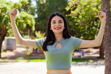 Young brunette woman wearing turquoise tee and orange short on city park, outdoors showing arms muscles smiling proud. Fitness concept. Look at my muscles!