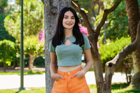 Young brunette woman wearing turquoise tee and orange short on city park, outdoors woman holding hands in pockets and looking at the camera pose for picture while leaning against tree.