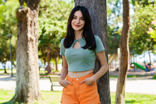 Cute caucasian woman wearing turquoise tee on city park, outdoors woman holding hands in pockets and looking at the camera pose for picture while leaning against tree.