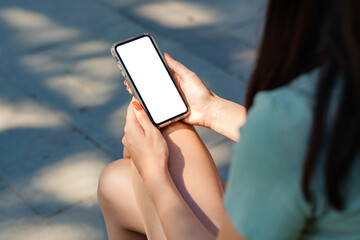Beautiful brunette woman wearing turquoise t-shirt standing on city park, outdoors hands holding phone touching finger mockup white blank display, mobile app tech concept, over shoulder closeup view.