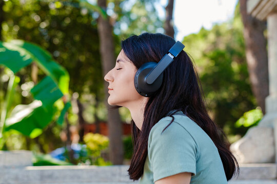 Side view of cute millennial woman wearing turquoise tee and orange short on city park, outdoors listening music in headphones and eyes closed. Enjoy good quality sound, stress free concept.