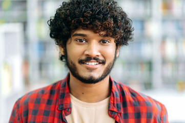 Close-up photo of curly-haired confident positive mixed race guy, Indian or Arabian male, in casual wear, manager, designer, student or freelancer, stands indoors, looks at camera, smiling friendly