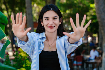 Cheerful brunette woman wearing blue striped shirt on city park, outdoors showing and pointing up...