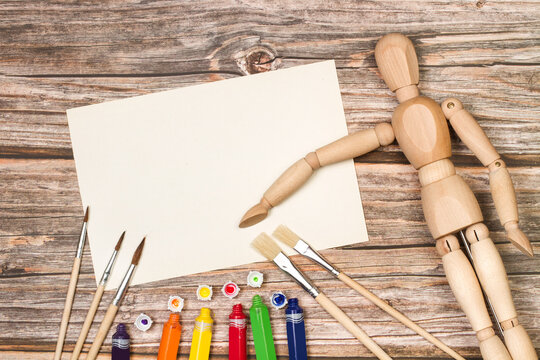 Pinceles y pinturas artisticas junto a un papel en blanco para pintar y un mu&ntilde;eco de madera articulado sobre un fondo de madera r&uacute;stico. Vista superior y de cerca. Copy space