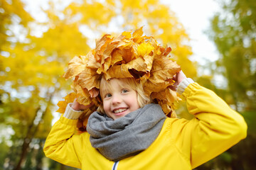 Cute little boy with a wreath of maple leaves on his head on sunny autumn day. Child having fun during stroll in the forest. Active outdoors leisure for children - obrazy, fototapety, plakaty
