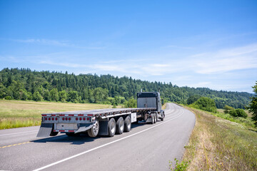Black classic big rig semi truck tractor transporting empty flat bed semi trailer running on the summer winding highway road in Columbia Gorge