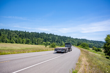 Classic black big rig semi truck with empty flat bed semi trailer driving on the winding summer...
