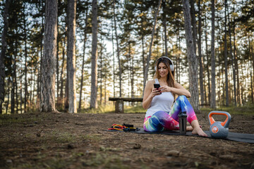 One woman adult caucasian female using mobile phone while taking a brake during training in the forest woods or park in summer day with kettlebell equipment listen music on earphones and mobile app