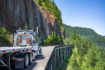 Pro classic black big rig semi truck with empty flat bed semi trailer going to warehouse for the next load running on the bridge around the rock cliff on mountain road in Columbia Gorge