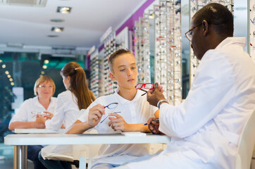 Teenage boy looking for stylish glasses with qualified African-American optician in optical shop