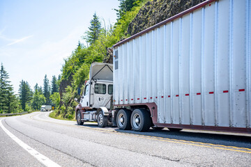 Two big rigs semi trucks with different semi trailers running towards each other on the winding mountain road with cliff and trees