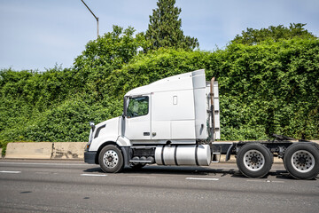 White big rig semi truck with truck driver cab sleep compartment driving on the wide highway road to warehouse for pick up loaded semi trailer