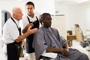 Skilled elderly hairdresser controlling haircutting of African-American male client performed by...