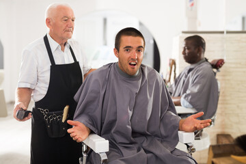 Young man unpleasantly surprised by haircut performed by elderly hairdresser at barber shop.