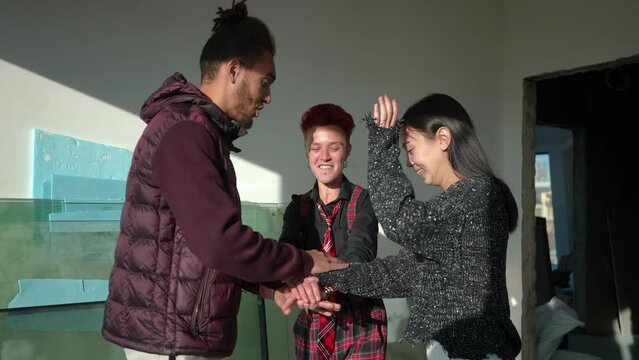 Joyful friends stacking hands in slow motion laughing gesturing thumbs up. Happy relaxed African American Asian and Caucasian man and women having fun meeting in building under construction