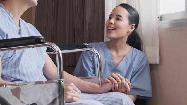 Uniformed young Asian female therapy doctor holding hands wheelchair male patient at window to support and motivate recovery, cancer illness after chemo medical treatment in hospital inpatient room.