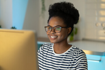 Cheerful joyful black girl remote worker wearing eyeglasses looking at computer monitor while...