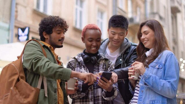 Close up portrait of mixed-race young people standing on street in city with coffee looking at smartphone screen and talking choosing place to go. Friends males and females walking in town, tourism
