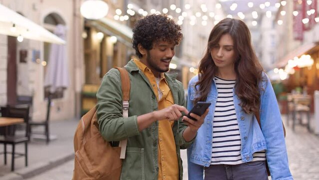 Portrait of cheerful nice couple looking at smartphone texting and surfing internet outdoors in town. Mixed-race handsome man and beautiful Caucasian woman talking in decorated street, couple concept