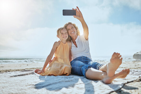 Carefree mother and daughter taking a selfie while sitting on the beach. Happy little girl and grandmother smiling while taking a picture on a cellphone while on holiday. Mom and daughter bonding