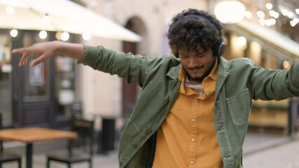 Camera approaching young happy handsome man dancing on street with many restaurants in good mood. Mixed-race stylish cheerful guy listening to music in headphones enjoying and making funny moves