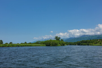 The bed of the blue river bends between the green banks. A picturesque mountain range against the background of azure sky and clouds. Copy space. Kamchatka. River Bystraya