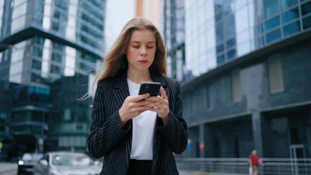 Businesswoman Ordering Taxi Via Smartphone App Standing Outdoor. A Young Business Woman Is Waiting For A Taxi And Looking In The App On Her Phone While Standing In The City Among The Skyscrapers.