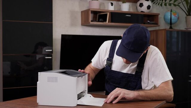 Close-up of a handyman in overalls, a white t-shirt and a cap with a screwdriver in his hands repairs the printer in the office or at the client's home. Call a home repairman.