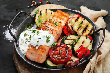 Salmon steak with vegetables and white sauce on dark background.