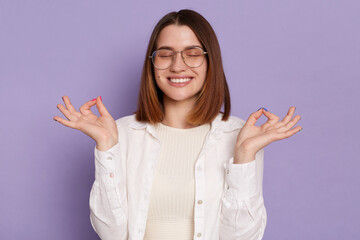 Image of positive satisfied delighted young adult Caucasian woman wearing white shirt, practicing yoga, relaxing, keeps eyes closed, posing isolated over purple background.